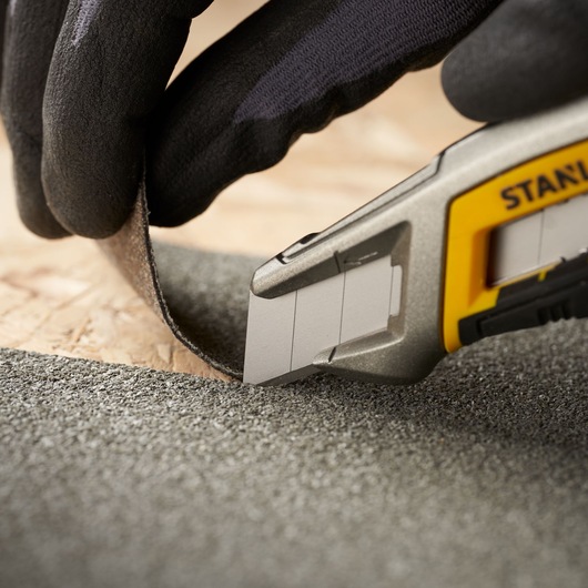 STANLEY 3-11-718 utility knife cutting abrasive material, close-up photo showing blade and gloved hand.