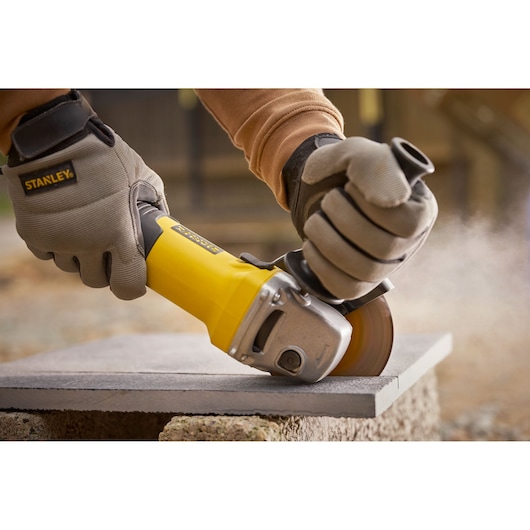Photo of STANLEY FMEG225VS grinder cutting stone slab outdoors, worker wearing protective gloves.