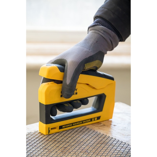 STANLEY FMHT0-80551 tacker in yellow and black, held by a gloved hand, pressing mesh onto wood surface.