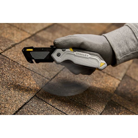 A close-up view of a hand wearing a grey glove holding a STANLEY folding utility knife, cutting into roofing shingles.