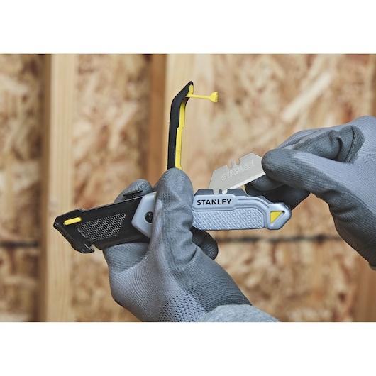 A pair of gloved hands replacing the blade of a Stanley utility knife, with a wooden wall background.