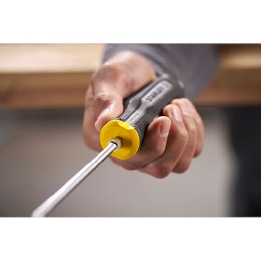Hand holding a Stanley screwdriver with a yellow and black handle, shown in use near a wooden surface.
