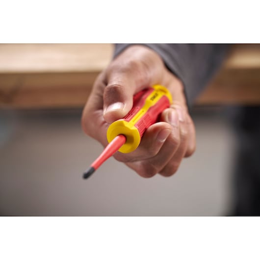 Hand holding a STANLEY insulated screwdriver with red and yellow handle near a wooden work surface.