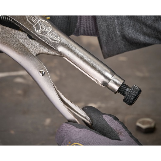 Close-up of a gloved hand adjusting a metal locking pliers tool with a screw on a workshop surface.
