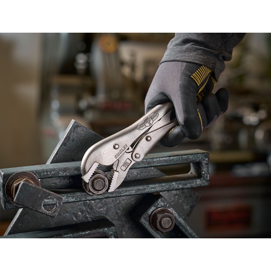Gloved hand uses a STANLEY locking pliers to grip a metal bolt in a workshop setting.