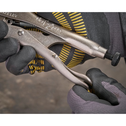 Gloved hands adjust a Stanley locking plier tool using the screw mechanism. Close-up detail of tool in use.