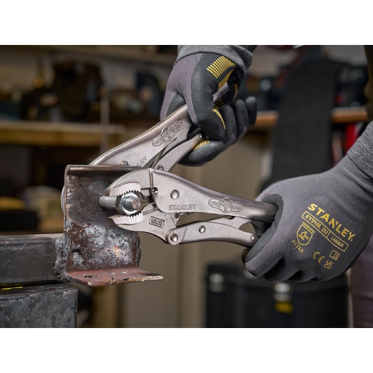 Stanley locking pliers held by gloved hands gripping a metal piece in a workshop setting.