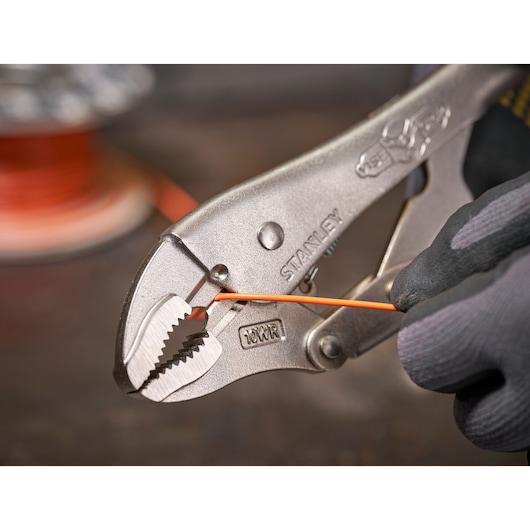 Stanley locking pliers gripping an orange wire, with gloved hand adjusting tool on a workbench.