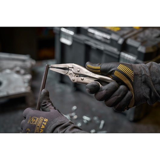 Gloved hands grip metal with Stanley locking pliers in a workshop setting, parts and toolbox in background.