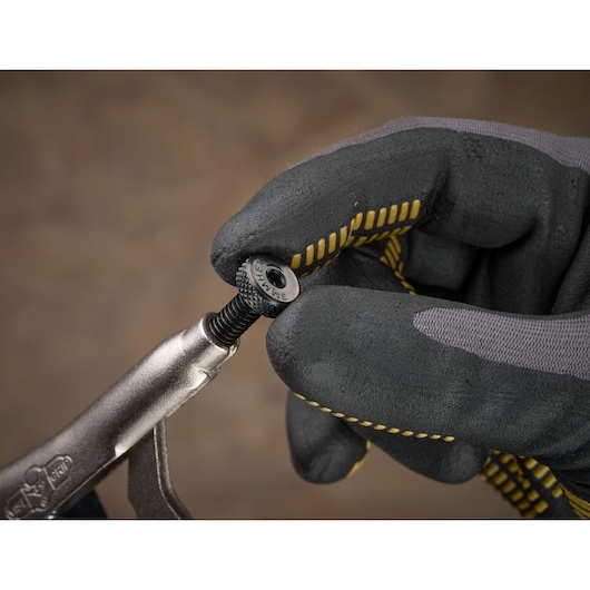 Gloved hand holding a metal screw with STANLEY tool against a brown background.