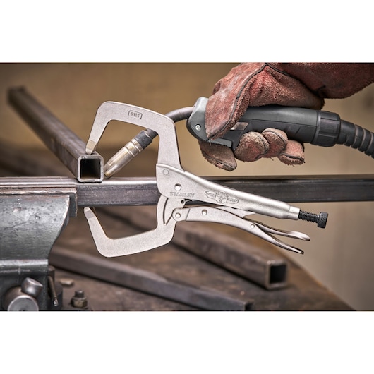 STANLEY locking C-clamp holding metal for welding, with a gloved hand using a tool on a metal workbench.