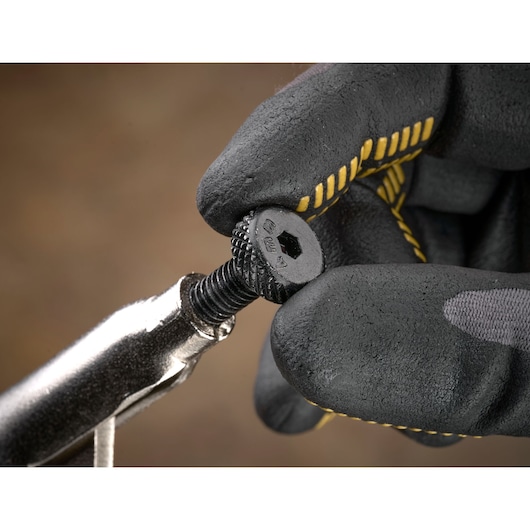 Gloved hand holding a textured screw with a wrench against a brown blurred background.