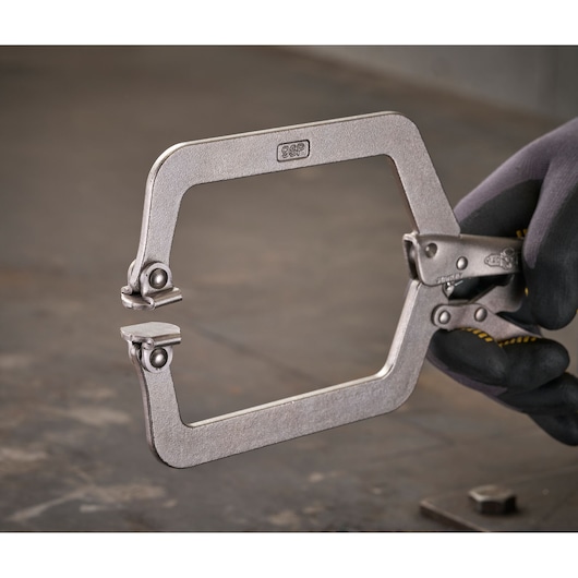 A metal locking C-clamp held by a gloved hand, used for securing materials in woodworking or metalworking tasks.