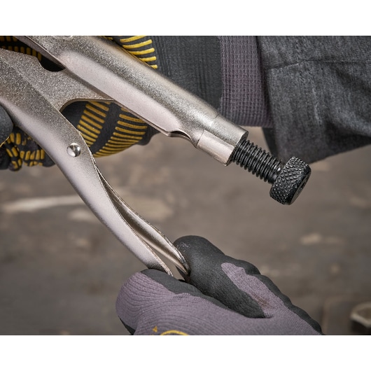 A gloved hand adjusts a metallic locking plier with a screw knob, shown in a workshop setting.