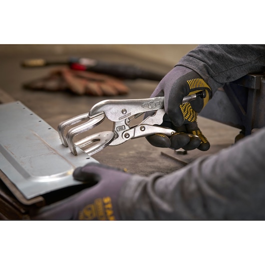 Person uses STANLEY locking pliers to hold a metal sheet on a workbench, wearing protective gloves.