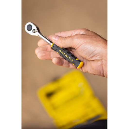 Photo of a hand holding a STANLEY FATMAX ratchet, SKU FMMT82683-0, black and yellow handle, blurred yellow toolkit below.