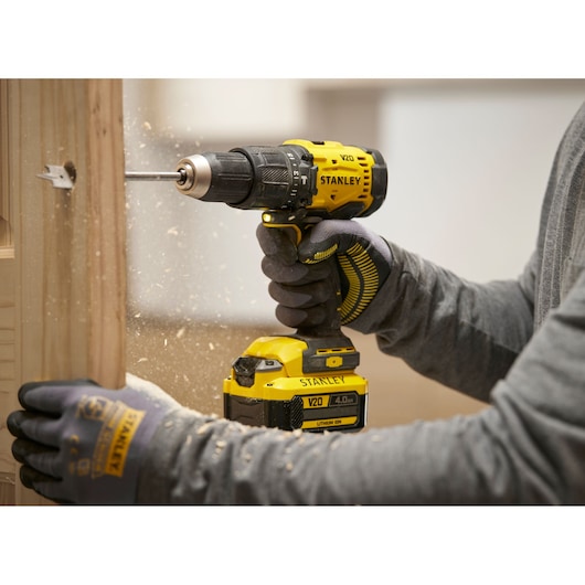 Person uses Stanley cordless drill to make a hole in wood, wearing gloves and grey shirt.