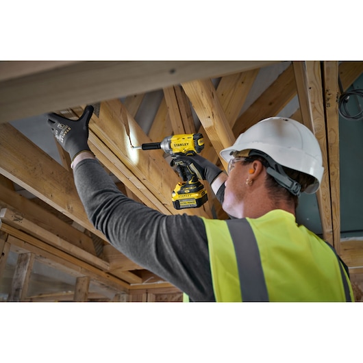 Worker using a STANLEY cordless drill on wooden beams, wearing gloves, helmet, and yellow vest at construction site.