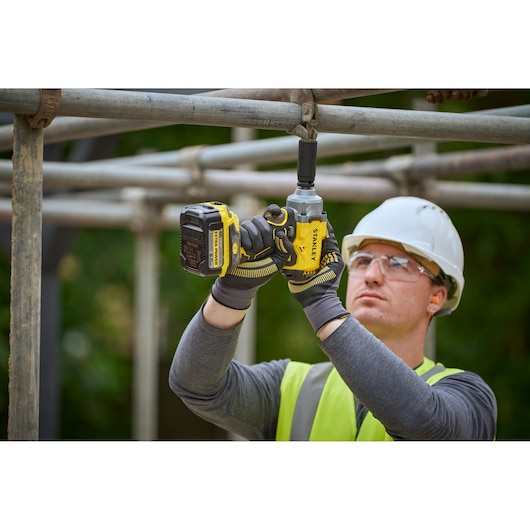 Worker uses STANLEY SFMCF921B cordless impact wrench on scaffolding, wearing safety gear and gloves.