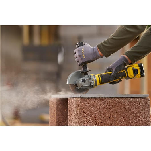STANLEY SFMCG700B cordless angle grinder in use cutting concrete block, close-up, shows dust and gloved hands.