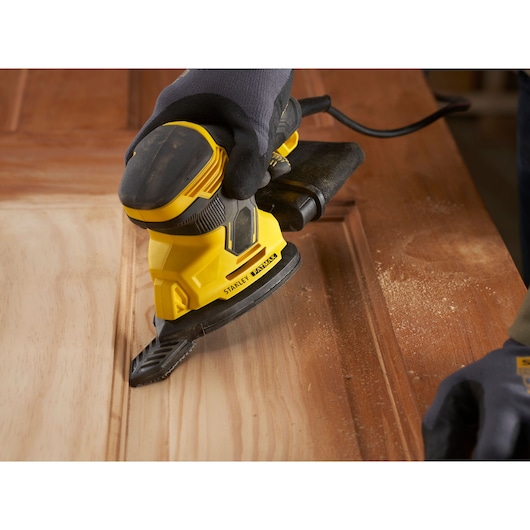 Photo of STANLEY SFMEW210S sander in use on wooden surface, close-up angle showing dust and ergonomic design.