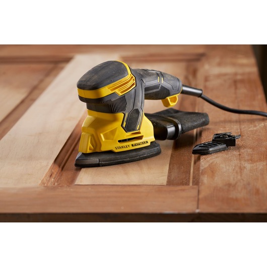 STANLEY SFMEW210S sander in use on wooden surface, close-up side angle, with attachments nearby.