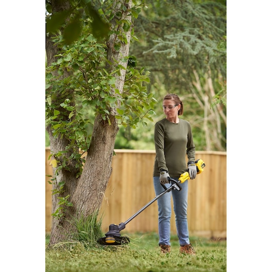 STANLEY SFMSTB930B grass trimmer in use outdoors, shown trimming grass at base of tree in a garden.