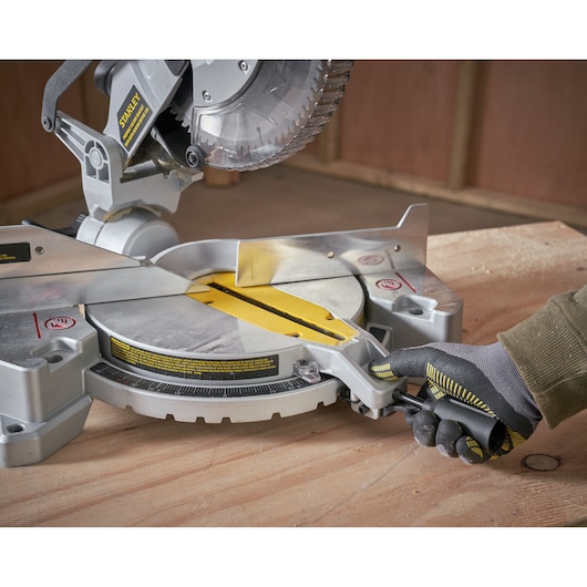 STANLEY SM1650 miter saw, close-up angle, silver and yellow details, glove adjusting saw on wooden surface.