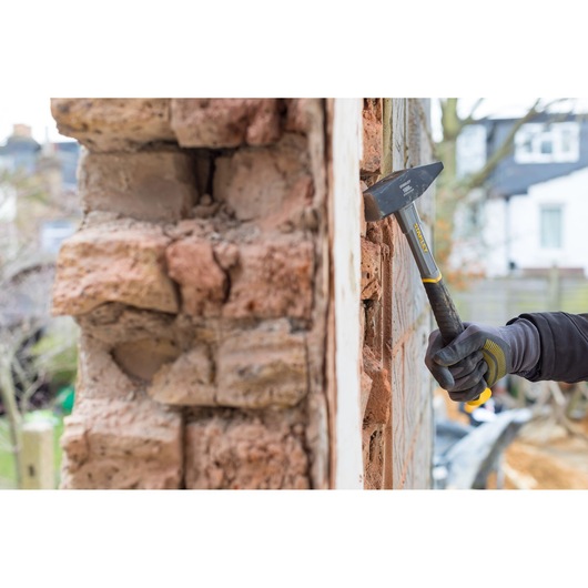 STANLEY STHT0-51910 brick hammer in use on masonry wall, close-up view, worker wearing gloves outdoors.