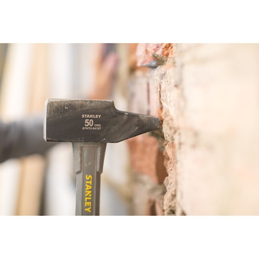 Stanley STHT0-54157 hammer in use against brick wall, close-up side angle, yellow branding visible.