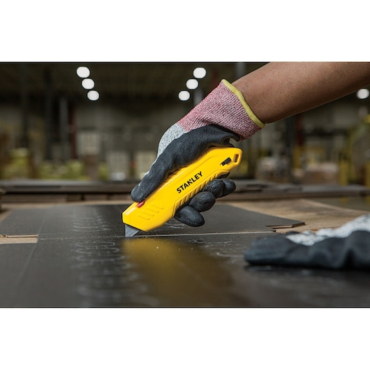 Photo of STANLEY STHT10368-0 yellow utility knife in a gloved hand cutting cardboard, photographed in a workshop.