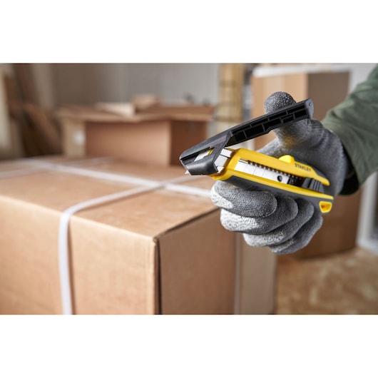 STANLEY STHT10501-0 utility knife held by gloved hand, cutting cardboard box, side angle.