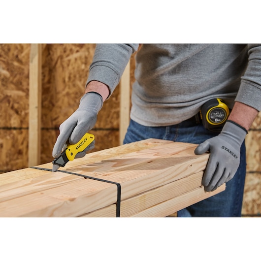 A person wearing gray gloves and a gray sweater uses a yellow STANLEY utility knife to cut a black strap securing bundled wooden planks. A STANLEY tape measure is attached to their belt. The scene takes place in a workshop with wooden walls.