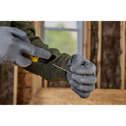 Close-up of a person wearing grey work gloves, inserting a STANLEY utility knife blade into a yellow utility knife handle at a construction site.