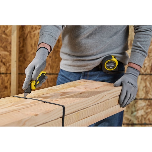 A person wearing grey gloves uses a yellow Stanley utility knife to cut a black strap around a bundle of wooden planks. A Stanley tape measure is clipped to the person's belt.