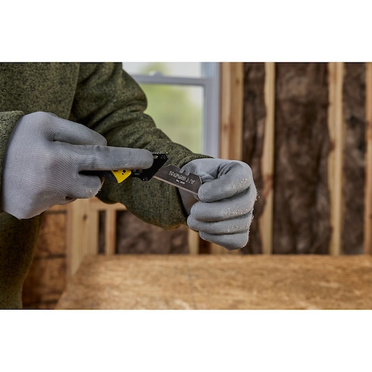 A person wearing gray work gloves and a green sweater is holding and adjusting a STANLEY utility knife in a workshop environment with wooden walls and insulation visible in the background.