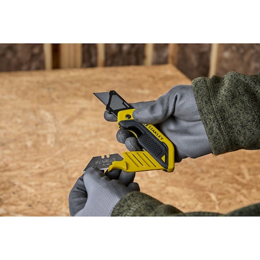 A person wearing gray gloves and a green sleeve is holding a yellow and black STANLEY utility knife, and replacing or inserting a razor blade into the tool. The background is a wooden work surface.