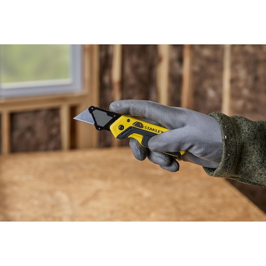 A gloved hand holding a STANLEY folding utility knife with a yellow and black handle, in a workshop setting.