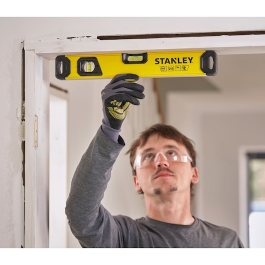 A person wearing a grey shirt and protective gloves is using a yellow STANLEY spirit level to check the alignment of a door frame in a home interior.