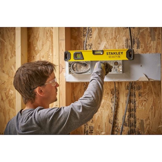 A person uses a STANLEY spirit level (model STHT41016-0) to check the alignment of a metal fixture on a wooden wall under construction.