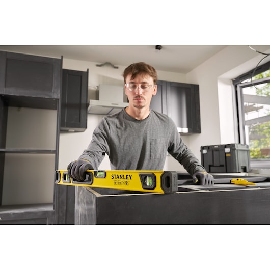 A person wearing gloves uses a yellow STANLEY spirit level to check the horizontal alignment of a kitchen countertop during installation. The scene is set in a modern kitchen under construction.