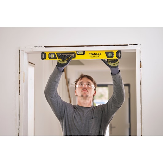 A person wearing gloves is holding a yellow Stanley spirit level against the top of a door frame in a room, checking alignment.