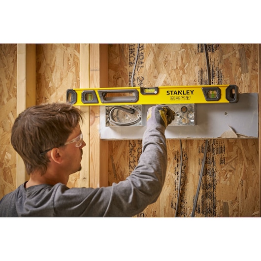 A person using a yellow STANLEY spirit level on a wooden wall frame during construction. The person's face is blurred for privacy. The spirit level is being used to align an electrical fixture.