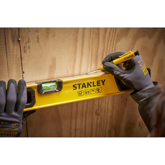 A person wearing work gloves uses a yellow STANLEY spirit level to mark a straight line on a wooden surface.
