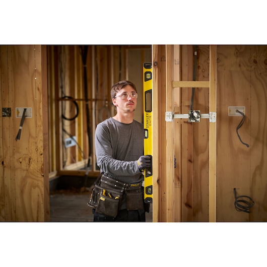 A person with a blurred face wearing a tool belt and gloves is using a yellow STANLEY spirit level to check vertical alignment on wooden framing inside a partially constructed building.