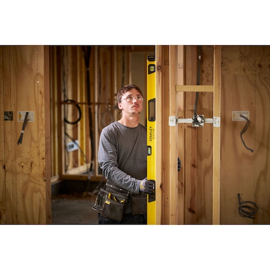 A person with their face blurred is holding a yellow STANLEY spirit level inside a wooden frame of a house under construction. The person is wearing a tool belt and gloves, and electrical wiring is visible in the wall.