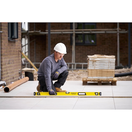 A person wearing a white hard hat and gloves is using a yellow Stanley spirit level to check the alignment of floor tiles at a construction site.