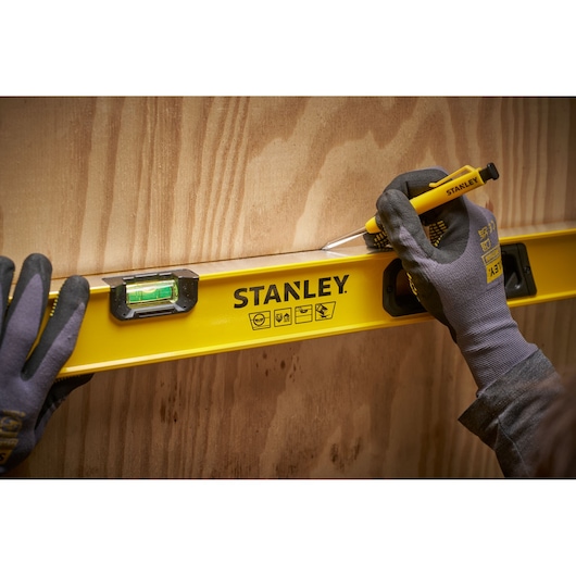 A person wearing gloves uses a yellow STANLEY spirit level and a pencil to mark a line on a wooden surface.