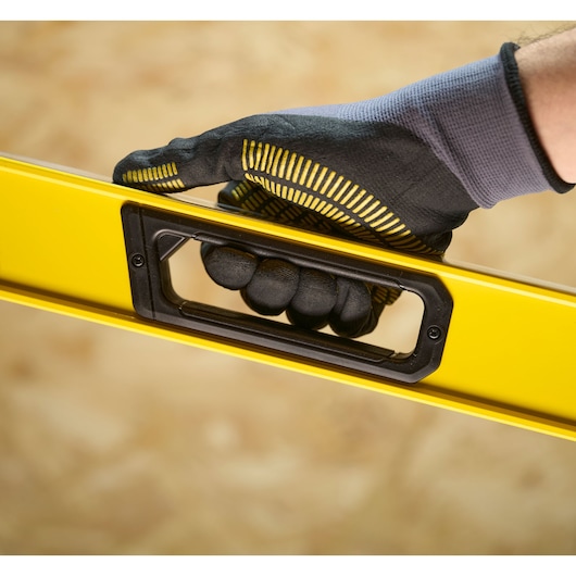 A close-up of a gloved hand holding a yellow Stanley spirit level by its built-in handle.