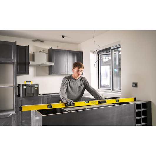 A person is using a yellow STANLEY spirit level to check the alignment of a countertop in a modern kitchen under construction. Black cabinets and a toolbox are visible in the background. The person's face is blurred.